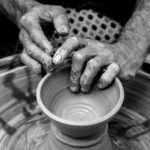 Two hands in black and white form a bowl out of clay on a potters wheel
