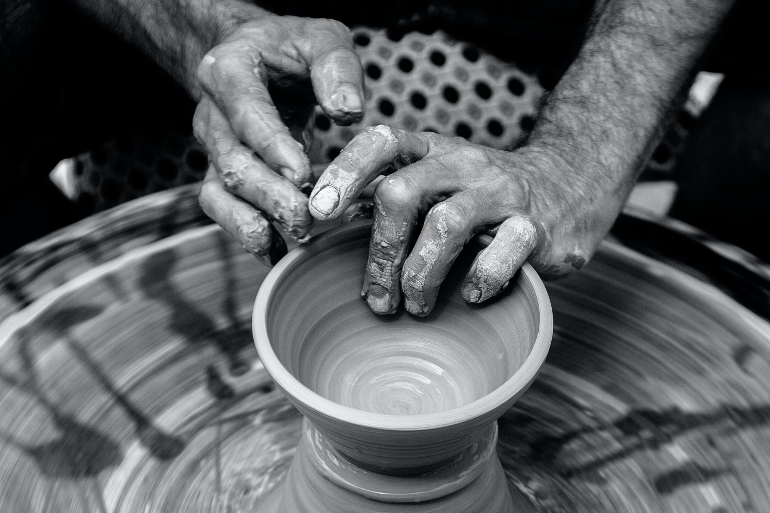 Two hands in black and white form a bowl out of clay on a potters wheel