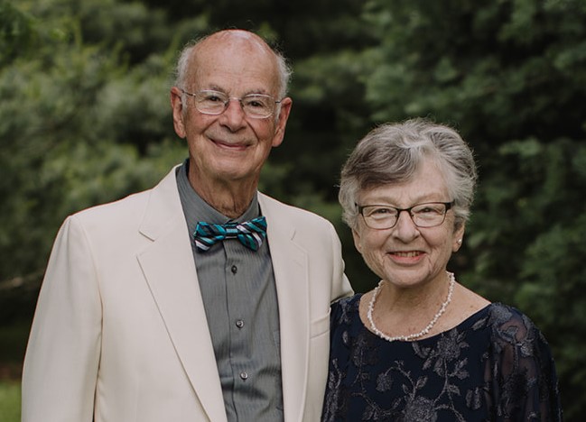 A man in his 70s stands with his arm around his wife who is shorter. He wars a white suit coat and bowtie, she has short hair, glasses, and wears a black dress.